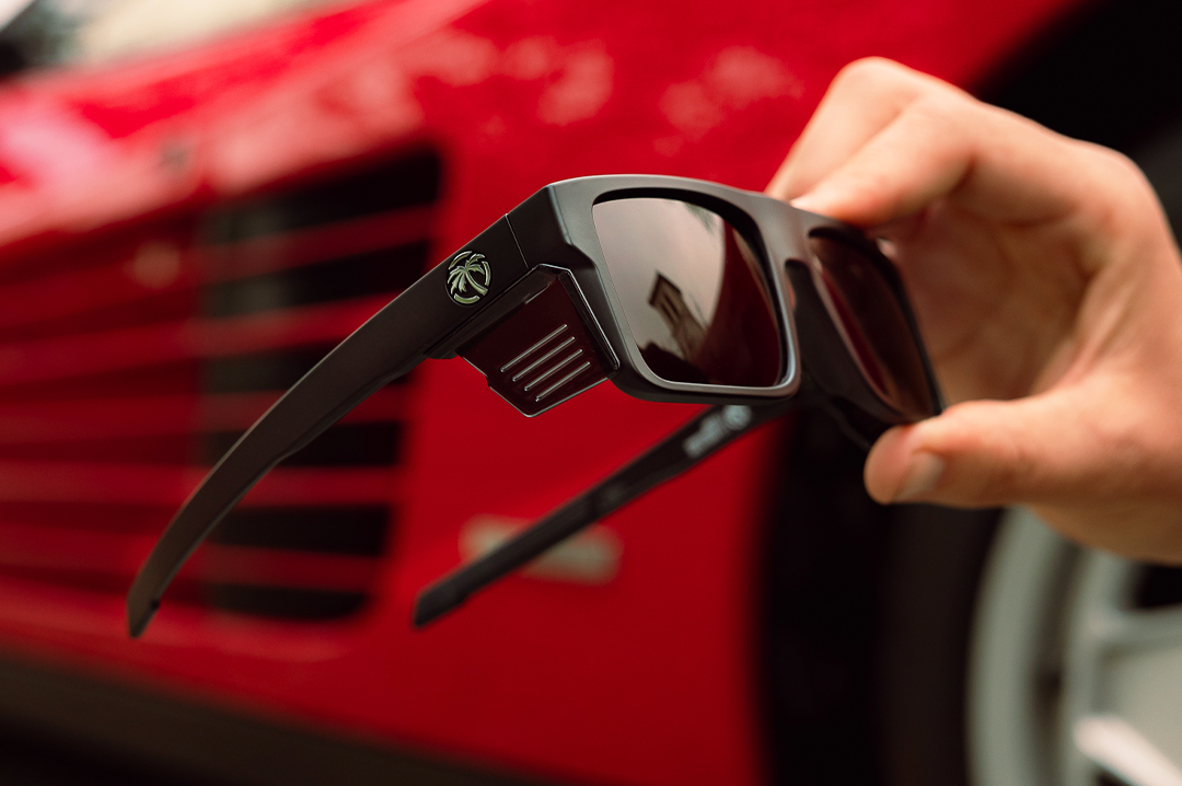 A hand holding up the Heat Wave Visual Performance Rayth Sunglasses with black frame, black lenses and matching side shields. 